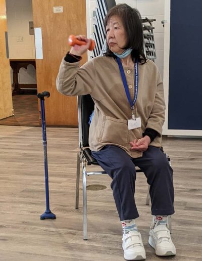 an elderly woman raises a hand weight while sitting in a chair, with her cane beside her chair
