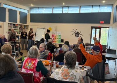 A group of people at a Halloween event with lots of tables. One man in an orange shirt holding up a big black fake spider.