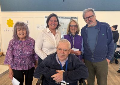Three women and two woman standing smiling. In front of them is a man sitting in a wheelchair, smiling.