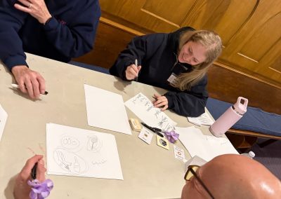 Two smiling older men sit at a table with a young woman, drawing pictures