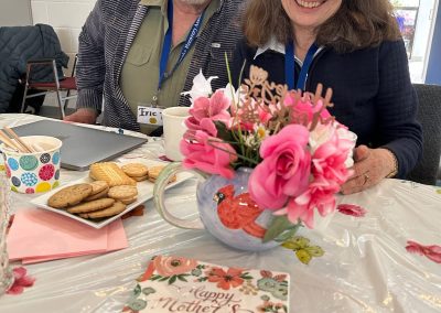 A seated man and woman smiling with a plate of cookies and a big teapot of flowers in front of them.