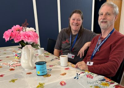 A smiling woman and man seated with cups of coffee, and a bouquet of big flowers in front of them