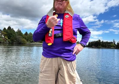 An older man in a purple shirt with a red lifejacket standing at the front of a dragon boat with a paddle lying on the strut in front of him.