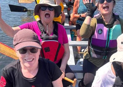 A group of women and men sitting in a dragon boat with their hands up, smiling.