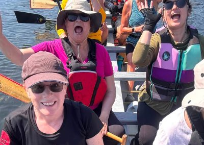 A group of women and men sitting in a dragon boat with their hands up, smiling.