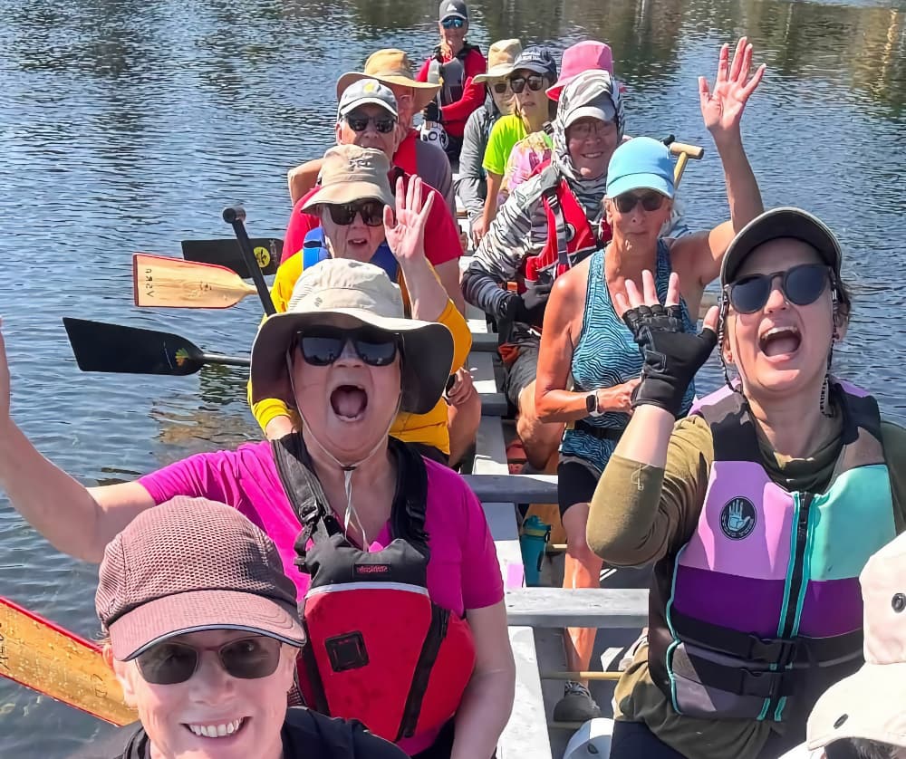dragon-boat-july2026-3-cropped A group of women and men in a dragon boat on rippling water smiling with their hands up.