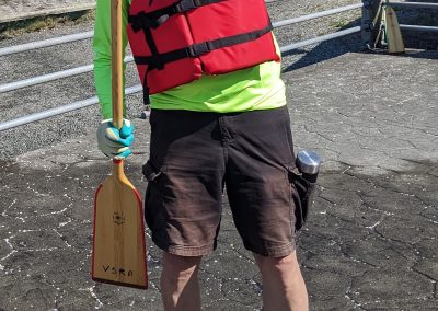 A man in a sunhat with a paddle in a bright UV shirt, shorts and lifejacket), showing off his paddling style.