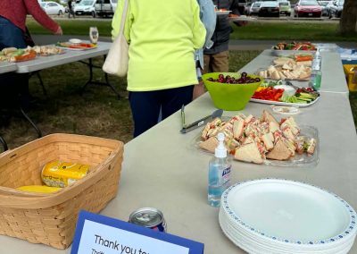 A long table outside in Victoria's Gyro Park with trays of sandwiches and veggies on it, some older people standing around.