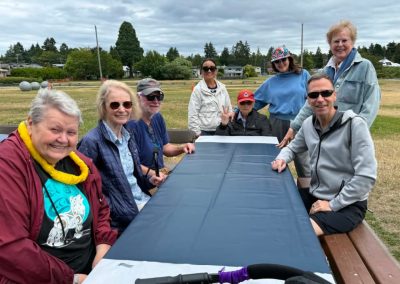 A group of adults sitting at an empty picnic table at a park with grey clouds but they're all smiling.
