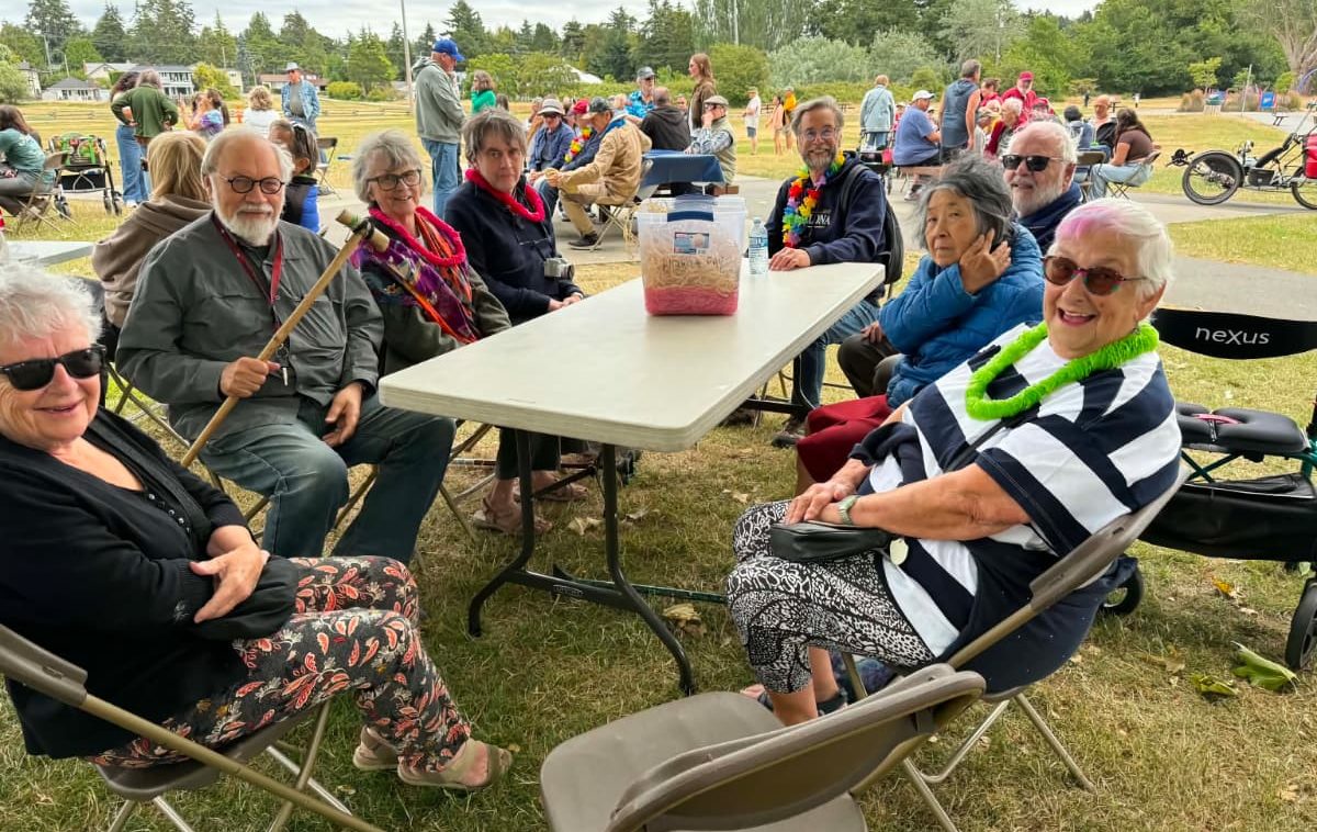 Eight smiling older people sitting around a picnic table, some of them wearing colourful leis. No food yet, but they look happy.