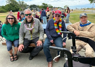 Three men and one woman sitting on a picnic table seat, one man with wearing a colourful lei (Hawaiian flowered necklace). Lots of people behind them, in a park on the ocean.